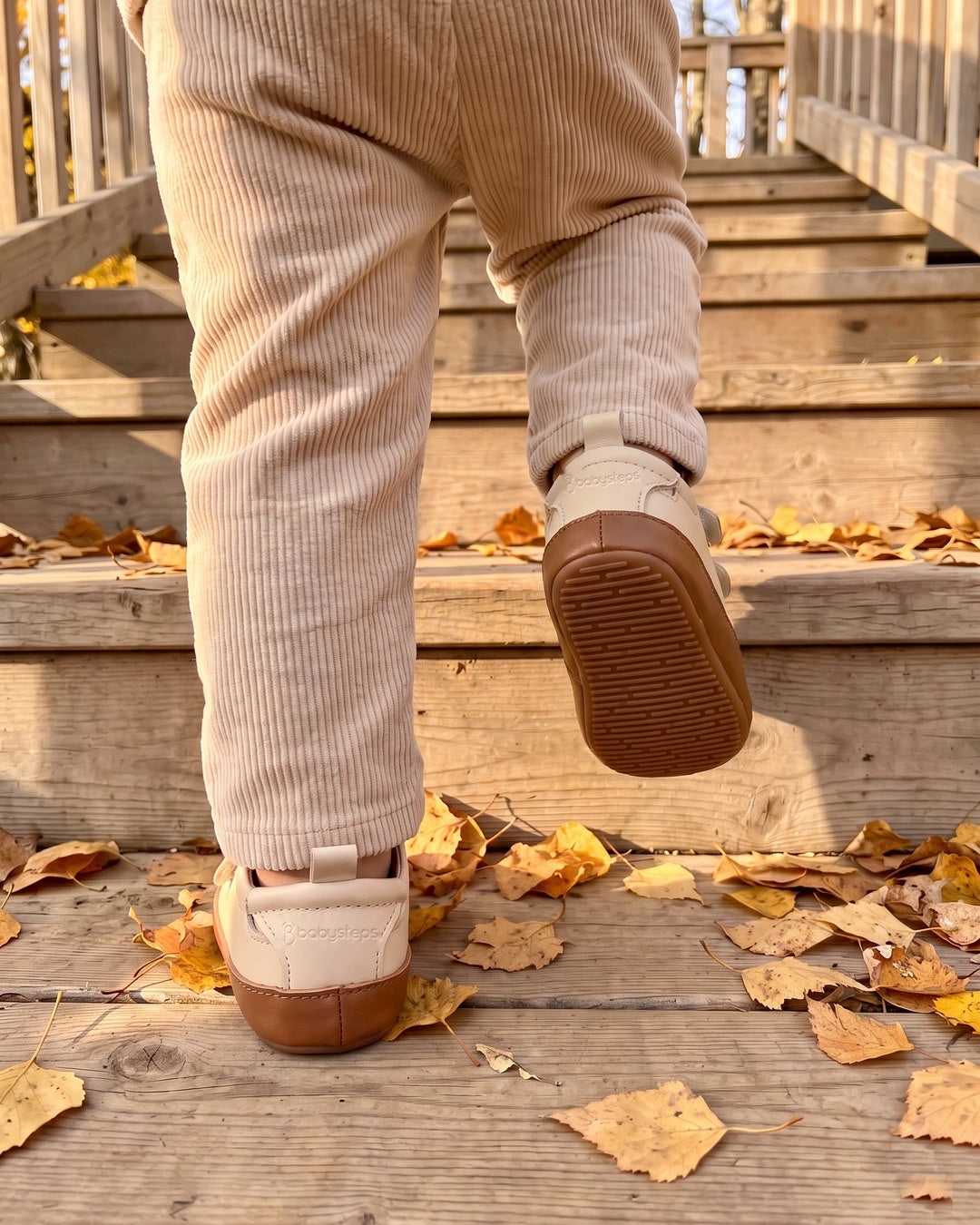 Barn i beige manchesterbyxor går uppför en trätrappa täckt av gula björklöv, i TabiSteps beige